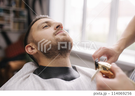 Professional hairdresser uses a hair clipper for fringing beard for a handsome man in barbershop. Side view Professional hairdresser uses a hair clipper for fringing beard for a handsome man in barbershop. Side view 114069256
