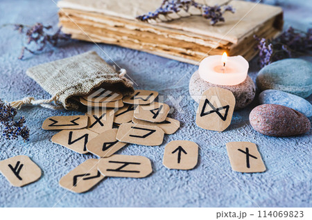 Fortune telling cardboard runes scattered from a bag, a candle, an old book and stones on the table 114069823