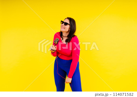Woman in sport clothes sings into a wired microphone and claps her hands on a yellow background Woman in sport clothes sings into a wired microphone and claps her hands on a yellow background 114071582