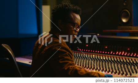 Portrait of african american audio technician uses mixing console with knobs and sliders in control room, editing recordings and adding sound effects. Skilled music producer at mixer. Camera A. Portrait of african american audio technician uses mixing console with knobs and sliders in control room, editing recordings and adding sound effects. Skilled music producer at mixer. Camera A. 114072523