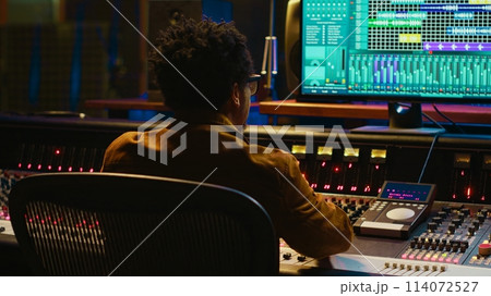 Audio expert counts down for artist singing in vocal booth, recording tracks to edit them after in post production. Producer operates mixing console and buttons or sliders, control room. Camera A. 114072527