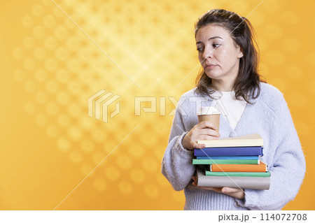 Portrait of woman with stack of books in hands holding disposable cup of coffee, isolated over studio background. Bookworm holding pile of novels and caffeinated drink early in the morning 114072708