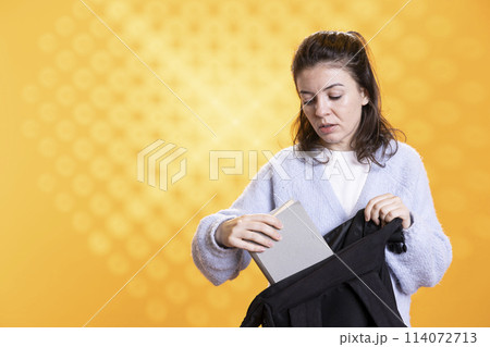 Portrait of woman rummaging through backpack, taking book out, isolated over studio background. Student removing textbook used for educational purposes from school rucksack Portrait of woman rummaging through backpack, taking book out, isolated over studio background. Student removing textbook used for educational purposes from school rucksack 114072713