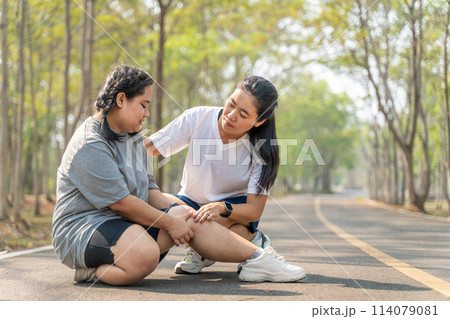 Overweight woman with knee injury sitting on the running track at a running park holding her painful knee while her female running friend sitting beside her in concern Overweight woman with knee injury sitting on the running track at a running park holding her painful knee while her female running friend sitting beside her in concern 114079081