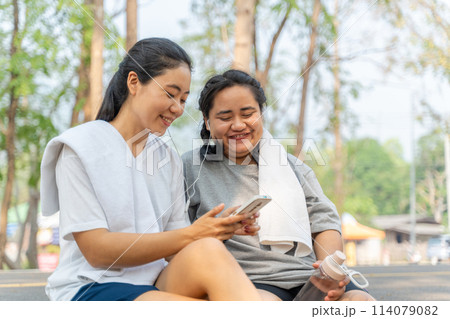 Two happy Asian female friends listening to music from a phone while sitting together at the curveside road after their morning run 114079082