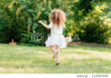 Easter egg hunt. Girl child Wearing Bunny Ears Running To Pick Up Egg In Garden. Easter tradition. Baby with basket full of colorful eggs. 114085994