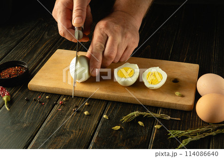 Hands with a knife cut an egg. Sliced eggs on a kitchen board for preparing a diet lunch with aromatic spices. Advertising space Hands with a knife cut an egg. Sliced eggs on a kitchen board for preparing a diet lunch with aromatic spices. Advertising space 114086640