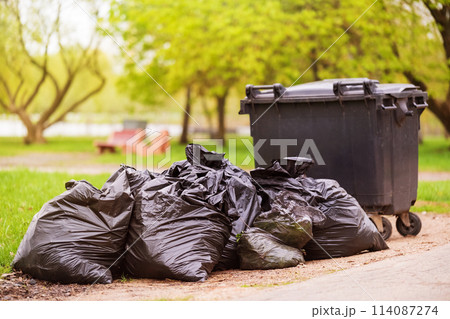 A pile of plastic garbage bags on the side of the road near a plastic container. Garbage bags on the street. Black trash bags. Concept of garbage collection, ecology, environmental cleaning 114087274