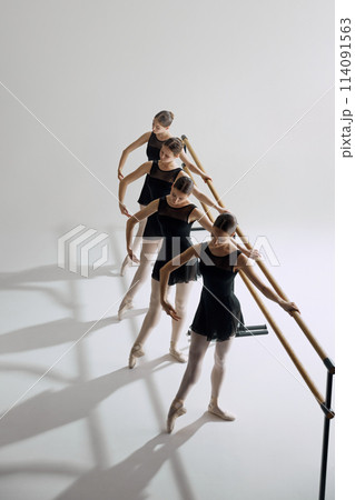 Four elegant girls, ballet dancers standing at barre and warming up, practicing against grey studio background. Focus on discipline 114091563