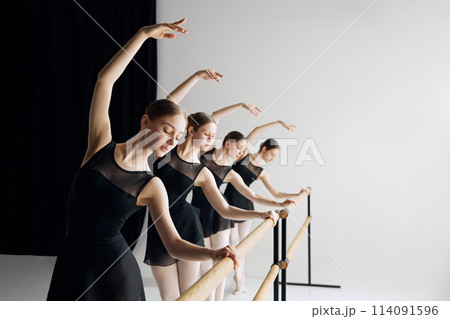 Beautiful talented teen girls, ballet dancers in black costumes standing at barre and practicing with focus and determination 114091596