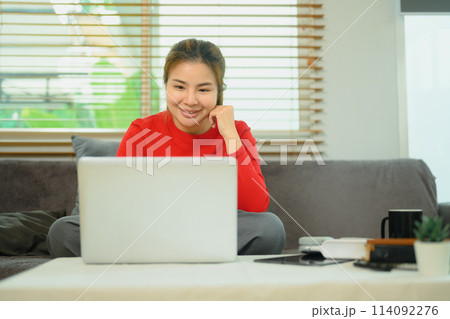 Cheerful young Asian woman sitting on couch working remotely at home on laptop Cheerful young Asian woman sitting on couch working remotely at home on laptop 114092276
