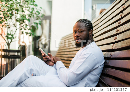 African american man in stylish ethnic dashiki wear viewing photos in social networks on mobile enjoying free time outdoors. Dark-skinned man watching funny video on smartphone and social networks 114101357