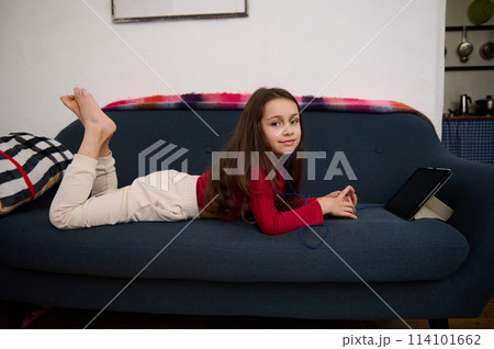 Caucasian cute elementary age school girl smiling looking at camera, lying on sofa, browsing websites, watching movies and having fun on her digital tablet with black mockup screen. Caucasian cute elementary age school girl smiling looking at camera, lying on sofa, browsing websites, watching movies and having fun on her digital tablet with black mockup screen. 114101662