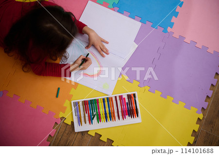 Little girl smiling drawing picture, lying on her belly on a multicolored puzzle carpet. People. Education. Back to school concept 114101688
