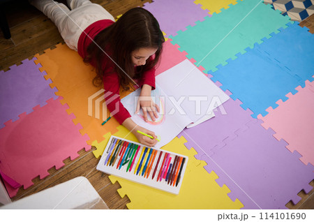 Top view of a talented little child, school girl taking out colorful pencil from pencil case, drawing beautiful cloud with rainbow, lying on a multi colored puzzle carpet in cozy home interior Top view of a talented little child, school girl taking out colorful pencil from pencil case, drawing beautiful cloud with rainbow, lying on a multi colored puzzle carpet in cozy home interior 114101690