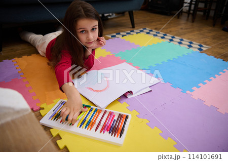 Little girl smiling looking at camera while taking a colorful pencil out of a box with pastel crayons, lying on her belly on a multicolored puzzle carpet. People. Education. Back to school concept 114101691