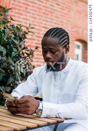 African american man in stylish ethnic dashiki wear viewing photos in social networks on mobile enjoying free time outdoors. Dark-skinned man watching funny video on smartphone and social networks 114102474