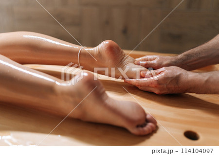 Male masseur massaging heel to relaxing woman lying on massage table in wellness center. Professional physiotherapist with strong hands performing foot massage. Concept of massage spa treatments. 114104097