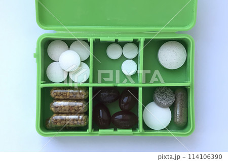 An open green fashionable pill container is lying on the white table. Many tablets and capsules are in the pillbox. Close-up An open green fashionable pill container is lying on the white table. Many tablets and capsules are in the pillbox. Close-up 114106390