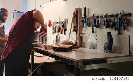 Woodworker in assembly shop using power drill to create holes for dowels in wooden board. Carpenter sinking screws into wooden surfaces with electric tool, doing precise drilling for seamless joinery Woodworker in assembly shop using power drill to create holes for dowels in wooden board. Carpenter sinking screws into wooden surfaces with electric tool, doing precise drilling for seamless joinery 114107143