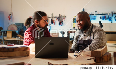 Cabinetmakers doing brainstorming while using CAD software on laptop, discussing wood designs. Carpenters in assembly shop discussing, using program on notebook to plan furniture assembling, camera B Cabinetmakers doing brainstorming while using CAD software on laptop, discussing wood designs. Carpenters in assembly shop discussing, using program on notebook to plan furniture assembling, camera B 114107145