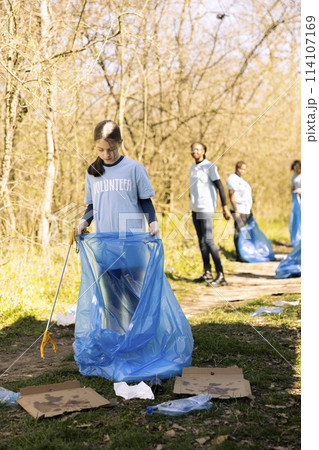 Community service volunteer reduces waste and cleans up forest setting, collecting garbage in blue trash bags. Young kid helps environment conservation by disposing of plastic waste. 114107169