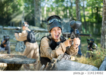 Excited girl paintball player aiming with paintball gun and shooting 114114082