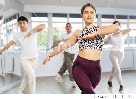 Happy girl and group of young active sport people practicing vogue dancing movements while training twist or charleston in dance hall 114114170