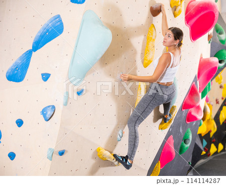 Young woman climbs steep artificial wall in sports complex and trains endurance Young woman climbs steep artificial wall in sports complex and trains endurance 114114287