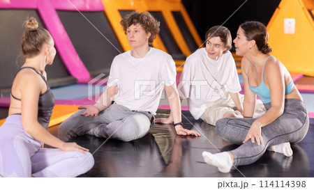Group of enthusiastic young people sitting on trampoline and discussing upcoming jumping workout in entertainment center 114114398