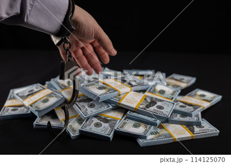 On a black background, a handcuffed hand reaches for a pile of dollar bills. A hand wants to steal a wad of dollars on a black background. Concept of money, dollars, theft. On a black background, a handcuffed hand reaches for a pile of dollar bills. A hand wants to steal a wad of dollars on a black background. Concept of money, dollars, theft. 114125070