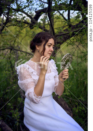 beautiful woman in white vintage dress with train and holding vintage mirror in spring garden at sunset 114125199