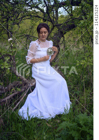 beautiful woman in white vintage dress with train and holding vintage mirror in spring garden at sunset 114125224