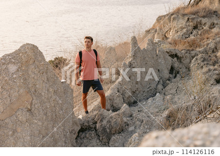 Handsome man in shorts with backpack on rocky ocean coastline with warm sunlight. 114126116