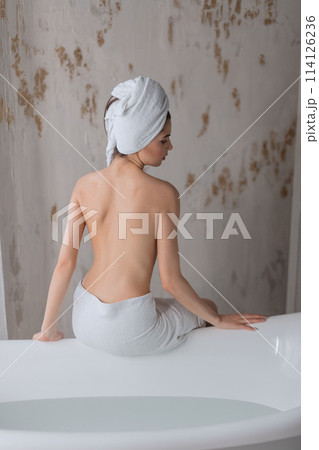 Young woman with naked back wearing towel on head, sitting on bathtub edge. 114126236