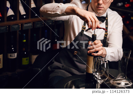 Bartender opening a wine bottle with corkscrew in restaurant with wine shelves. Bartender opening a wine bottle with corkscrew in restaurant with wine shelves. 114126373