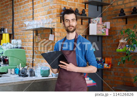 Young man in apron, food service worker, small business owner entrepreneur with work papers Young man in apron, food service worker, small business owner entrepreneur with work papers 114126826