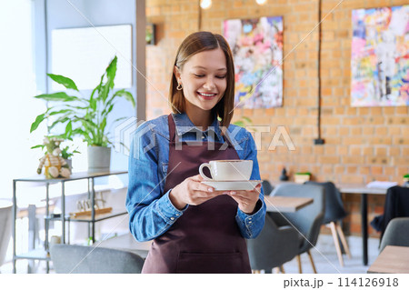Young woman worker, owner in apron holding cup of coffee in restaurant, coffee shop 114126918
