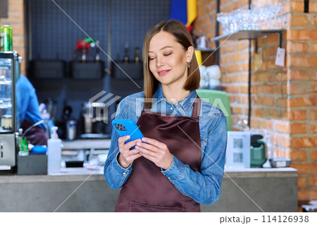 Young woman service worker in apron with smartphone in restaurant, coffee shop Young woman service worker in apron with smartphone in restaurant, coffee shop 114126938