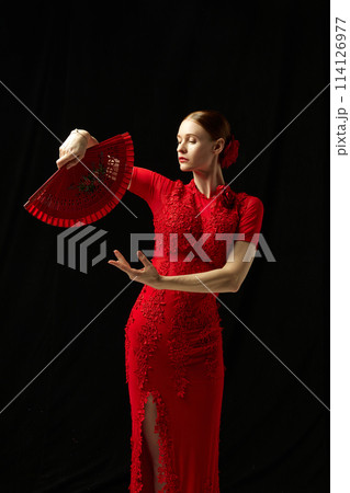 Flamenco female dancer in ornate red dress standing with elegance, posing with red fan against black background 114126977