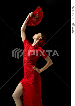 Flamenco female dancer in ornate red dress standing with elegance, posing with red fan against black background 114126978