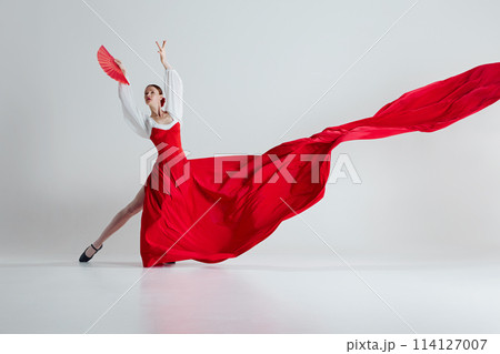In moment of pure flamenco artistry, female dancer in vibrant red dress and fan, performing flamenco with elegance against grey studio background In moment of pure flamenco artistry, female dancer in vibrant red dress and fan, performing flamenco with elegance against grey studio background 114127007