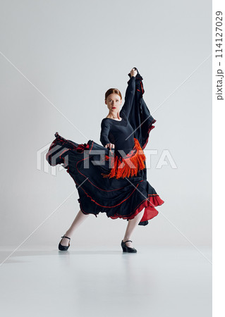 Spectacular performance of flamenco dance with elegant woman in red and black dress dancing with passion against grey studio background Spectacular performance of flamenco dance with elegant woman in red and black dress dancing with passion against grey studio background 114127029
