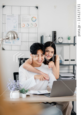 Cheerful young couple is seen enjoying a collaborative moment in their home office, with a focus on teamwork as they work on a laptop. Cheerful young couple is seen enjoying a collaborative moment in their home office, with a focus on teamwork as they work on a laptop. 114127131