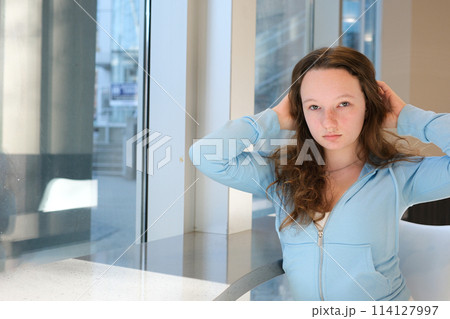 beautiful young girl teenager at table by window straightens hair looks into frames sports blue light jacket brown hair asian eyes white skin puffy lips european ukrainian young woman growing up 114127997