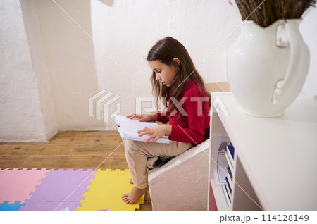 Side shot of Caucasian cute little child girl sitting barefoot on steps, enjoying reading a book encyclopedia fairytale in her cozy room. White bookshelf and vase of lavender flowers on the foreground Side shot of Caucasian cute little child girl sitting barefoot on steps, enjoying reading a book encyclopedia fairytale in her cozy room. White bookshelf and vase of lavender flowers on the foreground 114128149