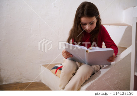 Caucasian cute little child girl sitting on steps at home, reading a book over white wall background. Literature. Erudition. Knowledge. World Book Day. Back to school concept. Copy advertising space 114128150
