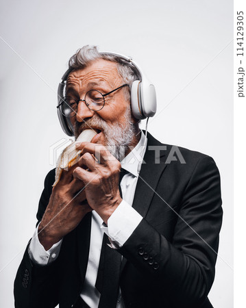 Mature businessman eating banana and listening to audiobook using headphones. Mature businessman eating banana and listening to audiobook using headphones. 114129305