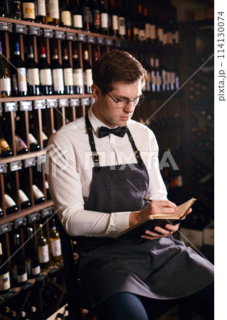 Wine steward looking at bottles of wine and writing in notebook at wine cellar 114130074