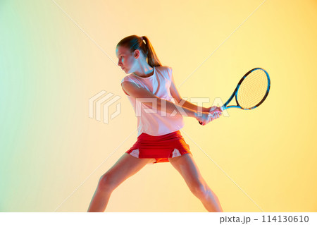 Dynamic portrait of sportive teen girl, tennis player in sports uniform training in action in neon light against blue-yellow background. Dynamic portrait of sportive teen girl, tennis player in sports uniform training in action in neon light against blue-yellow background. 114130610